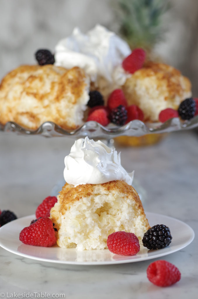 slice of pineapple angel food cake in bundt shape with whole cake behind it with cool whip and berries