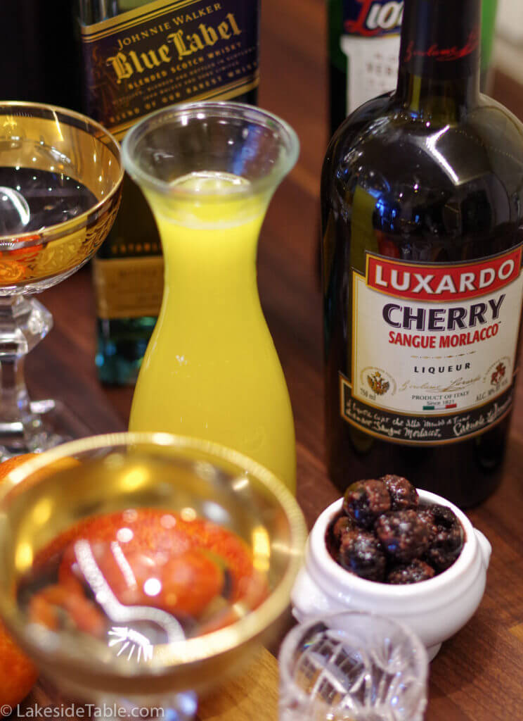decomposed ingredients for a blood and sand cocktail: cherries, blood orange juice, scotch and luxardo liqueur