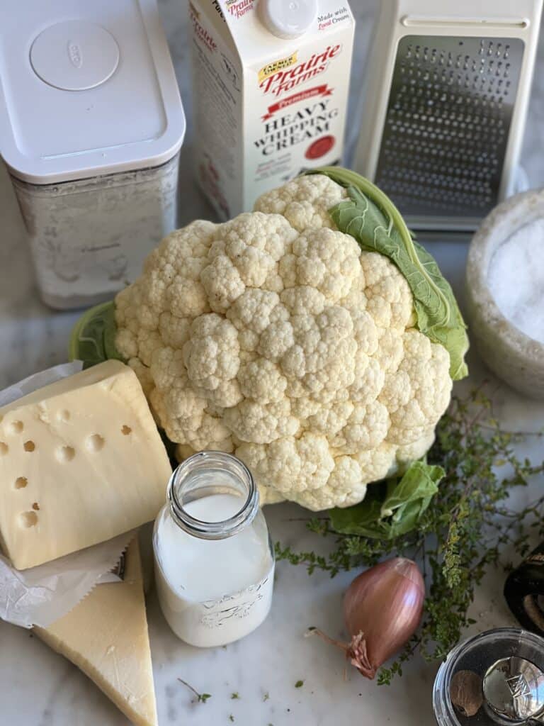 ingredients laid out for cauliflower gratin recipe: cauliflower, swiss cheese, parmasean cheese, cornstarch, salt, milk, heavy cream, nutmeg, shallot, and thyme 