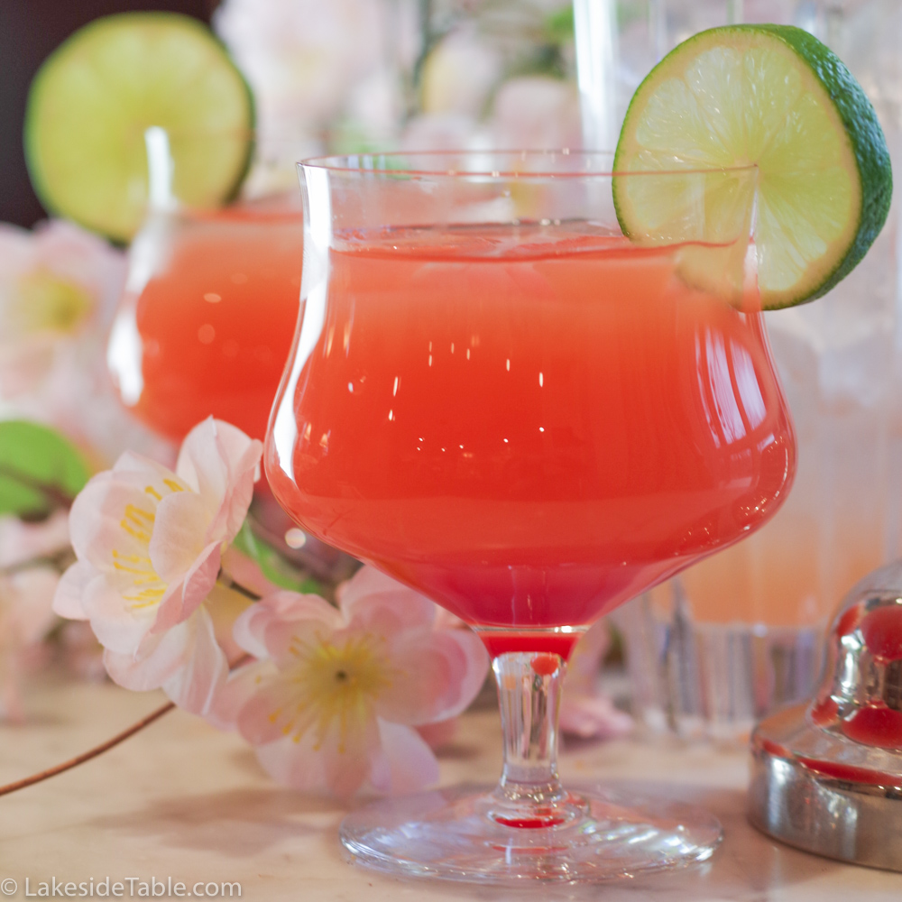 dark pink cocktail in a stemmed glass with a lime wheel on the side rim in front of blossoms