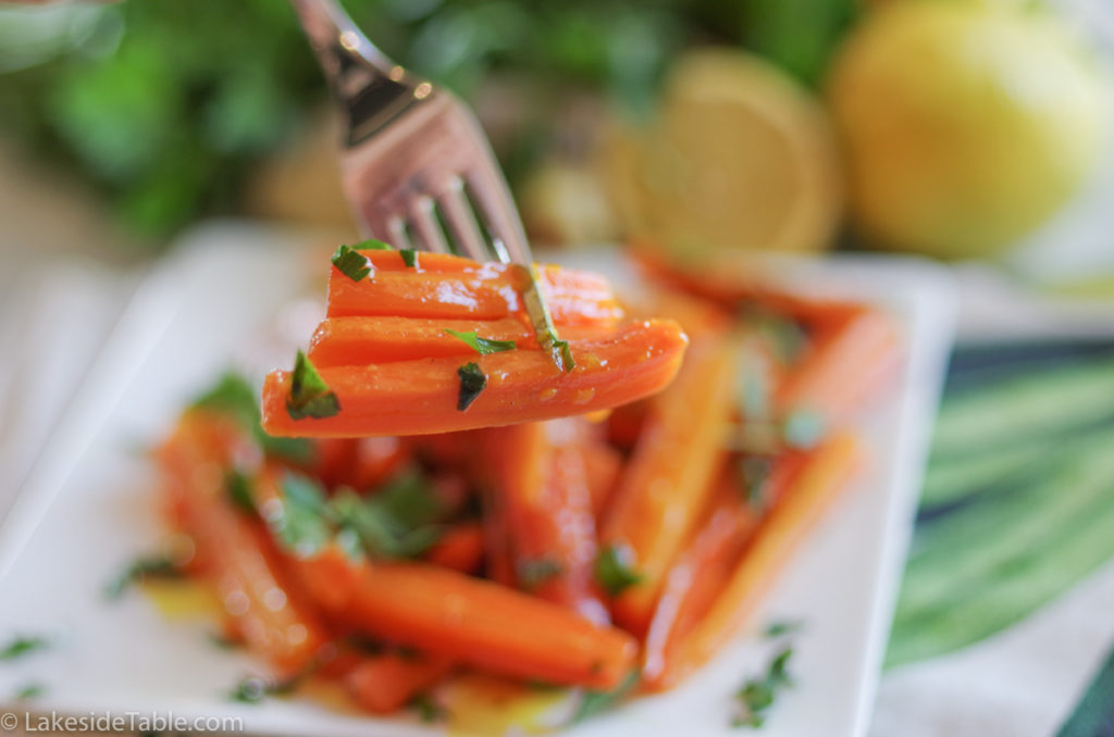 turmeric honey glazed carrots recipe - plate of carrots cut into long match sticks, sprinkled with parsley with a fork coming down into them. Behind this is an array of fresh parsley and lemons.