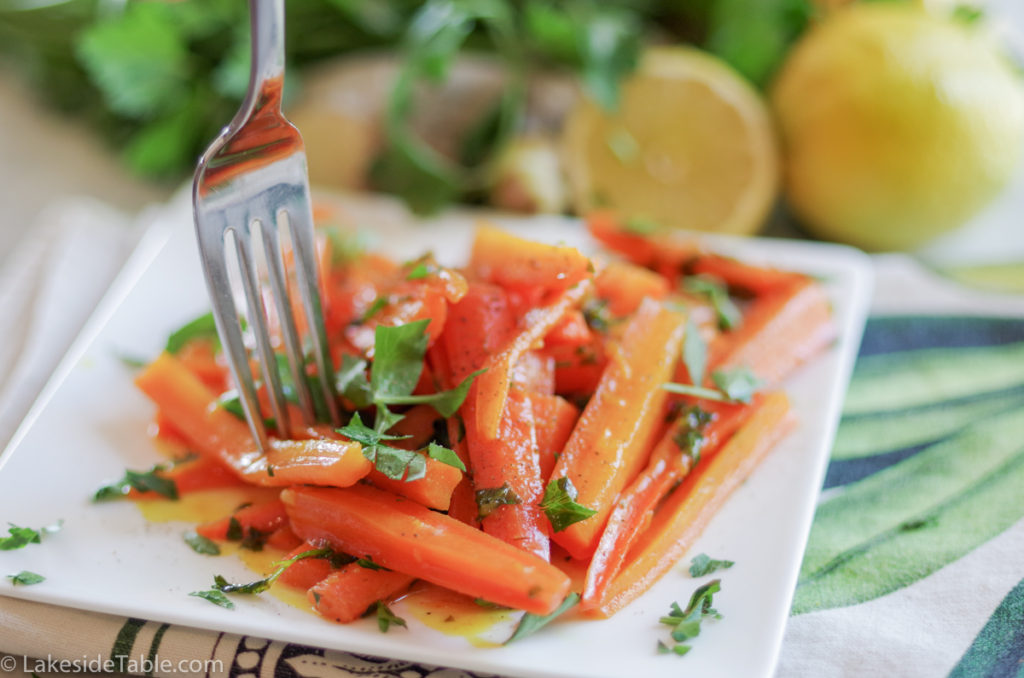 turmeric honey glazed carrot recipe - plate of carrots cut into long match sticks, sprinkled with parsley with a fork coming down into them. Behind this is an array of fresh parsley and lemons.