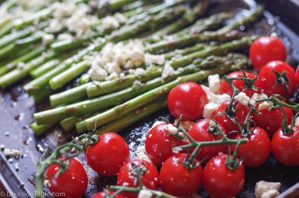 Bright green braised asparagus laid out on a baking sheet with white feta sprinkled on top in front of bright red cherry tomatoes