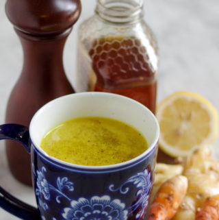 Hot mug of a yellow turmeric tonic with ice next to fresh turmeric root, ginger, lemon, honey and pepper