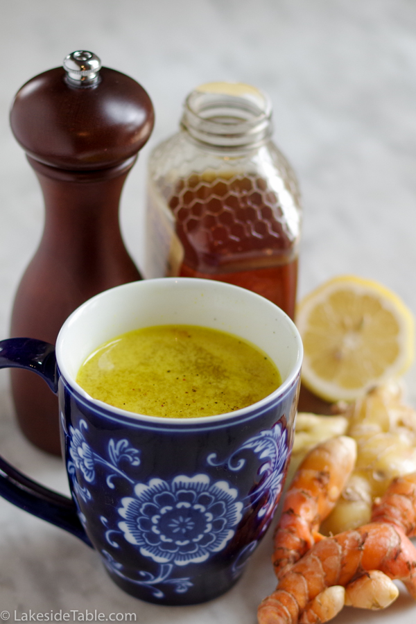 Hot mug of a yellow turmeric tonic with ice next to fresh turmeric root, ginger, lemon, honey and pepper