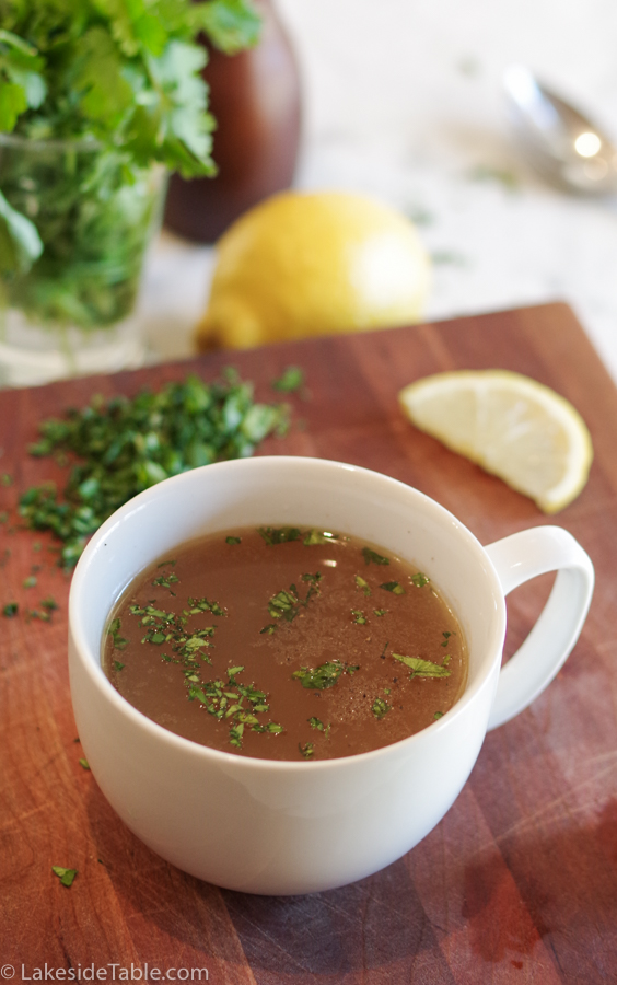 bowl of bone broth in white mug on butcher block in front of lemon wedge and green chopped parsley
