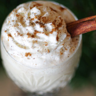 close up of a glass of homemade egg nog with whip cream and a cinnamon stick topped with ground nutmeg