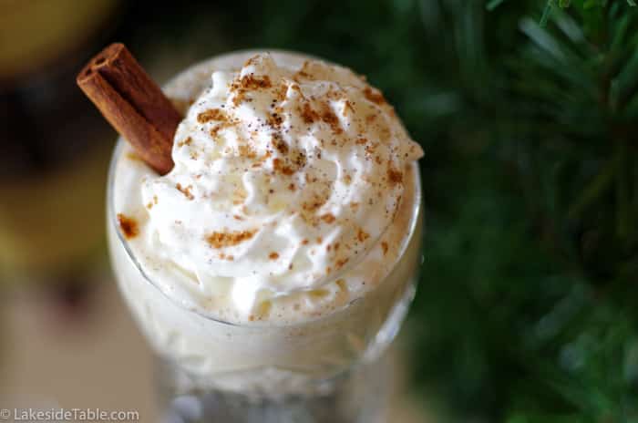 top down view of homemade egg nog with whip cream and a cinnamon stick topped with ground nutmeg