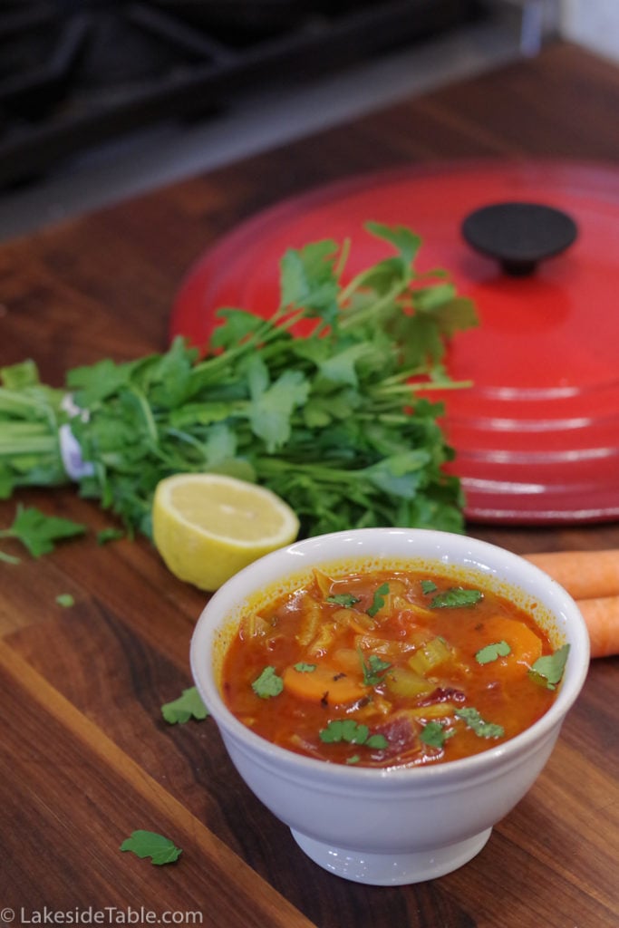 white bowl full of red cabbage soup with green parsley on top with lemon, parsley and a red lid in background