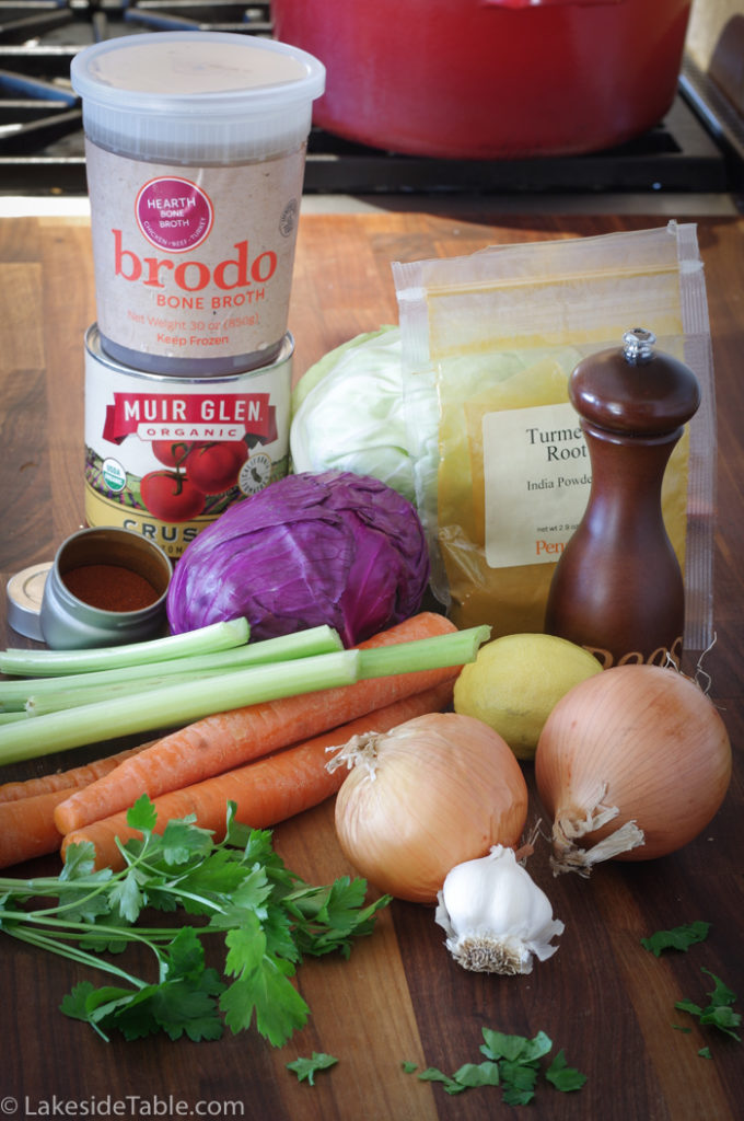 Detox Cabbage Soup ingredients laid out: brodo chicken broth, can of tomatoes, pepper, turmeric, cabbage, paprika, celery, carrots, lemon, onions, parsley and garlic