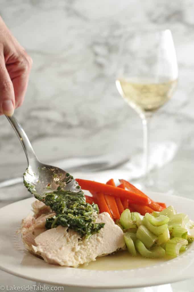 spooning bright green chimichurri sauce over poached chicken with a side of sauteed celery and glazed carrots with a glass of white wine in the background