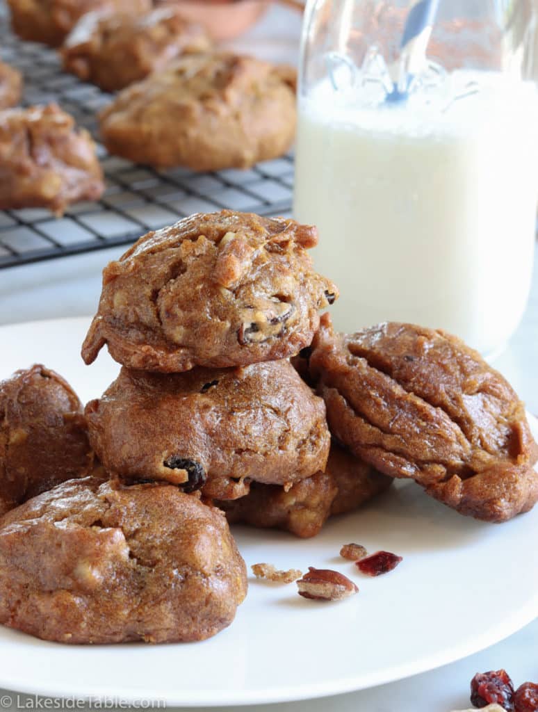 a stack of puffy persimmon cookies on a white plate in front of a glass of milk and cookies cooling on a rack