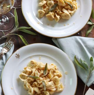 2 plates of finished tortellini pasta dripping with brown butter sauce topped with crispy sage leaves