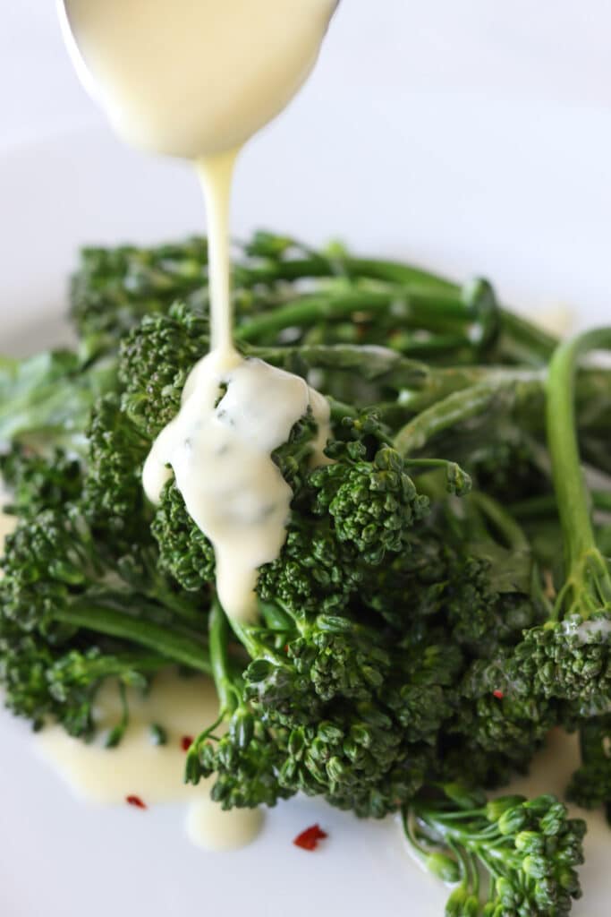 Light yellow sauce being slowly drizzled over bright green broccoli on a white plate