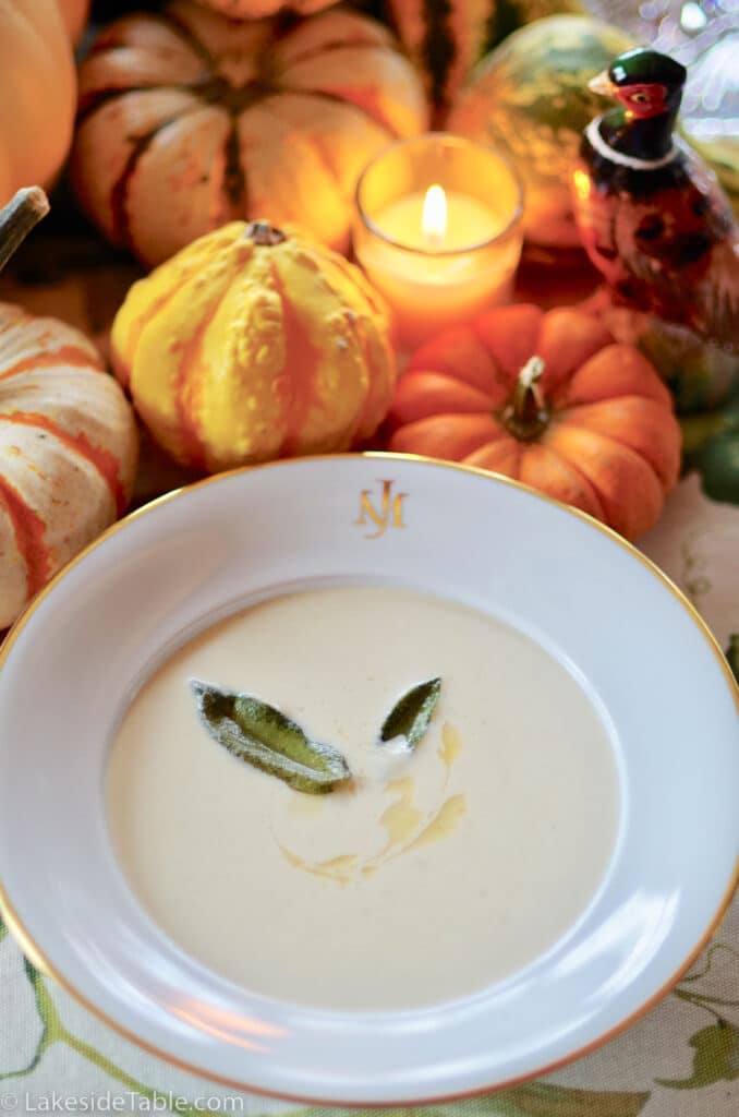 bowl of cream of cauliflower soup with sage leaves on top mini pumpkins behind bowl with a lit candle