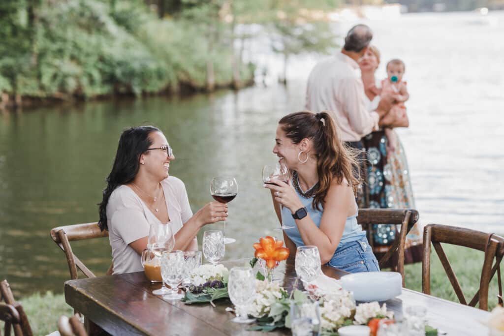 2 young women laughing at a table by a lake drinking wine
