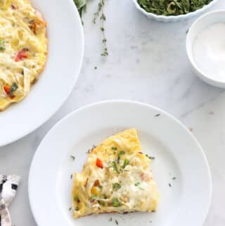 top down view of a slice of pasta frittata on a white plate on a white marble table scattered with herbs and a dish of salt
