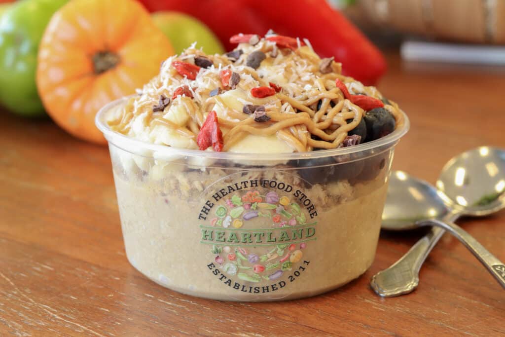 Pumpkin berry acai bowl in a clear 8 oz vessel with a Heartland Health Food sticker on the front topped with granola, blueberries, banana, and nut drizzle. Two spoons on the side.