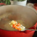 Steaming pot of soup behind a spoon full of onions, carrots, and potatoes