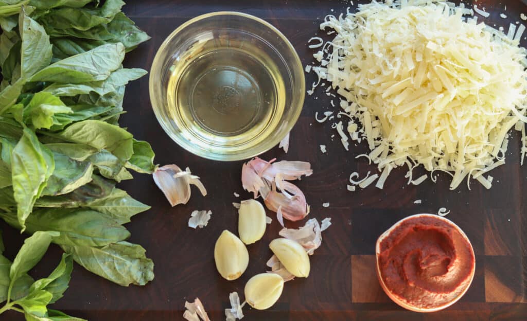 Pistou ingredients laid out on a cutting board: basil, oil, garlic, cheese, tomato paste