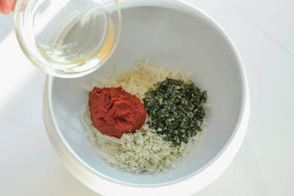 Pouring oil into pistou ingredients: cheese, chopped basil, garlic, and tomato paste