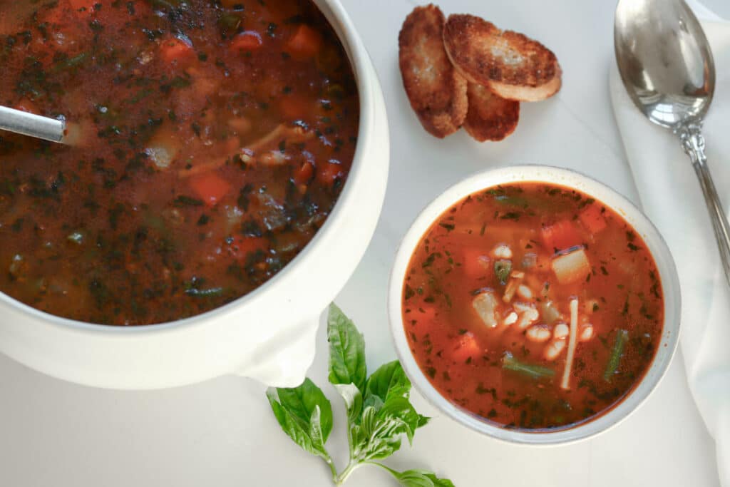 Large white Tureen of soup behind a white bowl of soupe au pistou with fresh basil, garlic, and french bread