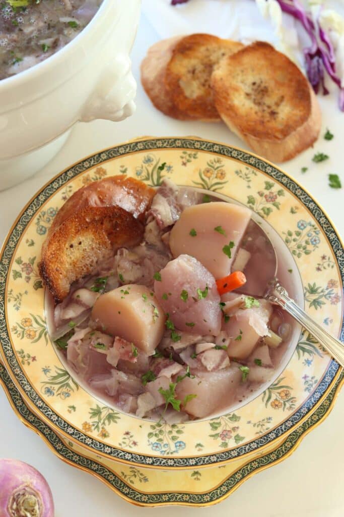 Purplish cabbage soup in a bowl with big chunks of potatoes, turnips, and carrots with a side of toasted French bread