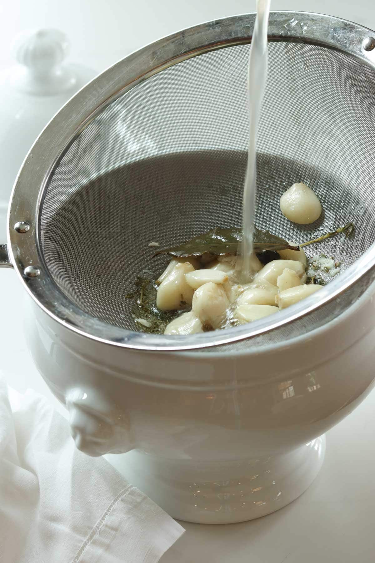 straining the garlic soup into a white soup tureen through a mesh sieve