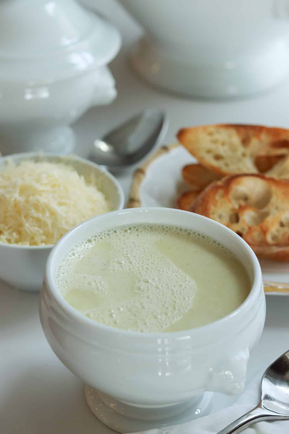 Julia Child's Aïgo Bouido soup in a white lion head soup bowl in front of a white soup tureen, toasted french bread, and grated cheese.