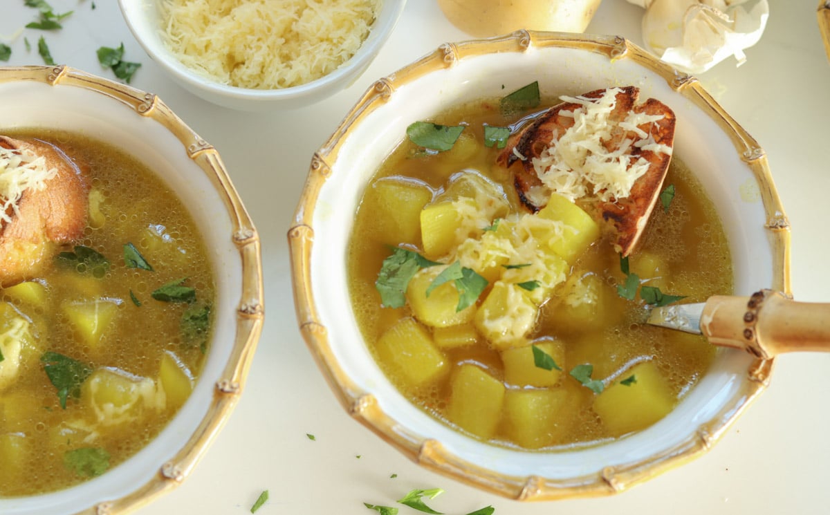 two bowls of garlic soup with saffron and potatoes in bamboo lined bowls on a white table top with parsley sprinkled around